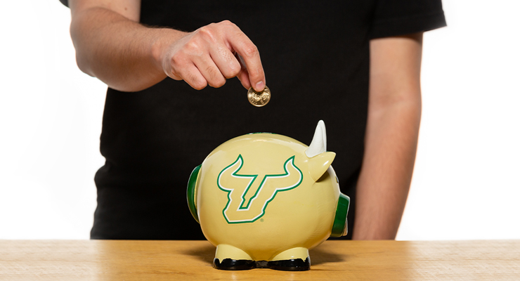 A person inserting a coin into a USF piggy bank.