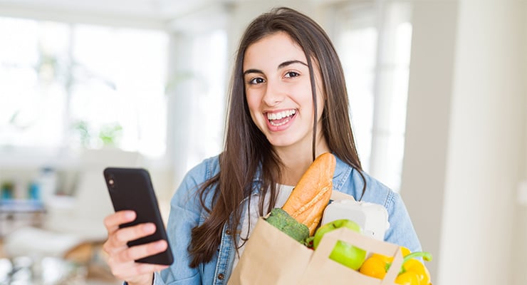 A student smiling while holding her phone and groceries.