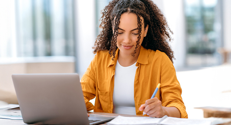 A woman sitting at a table taking notes and using her laptop.