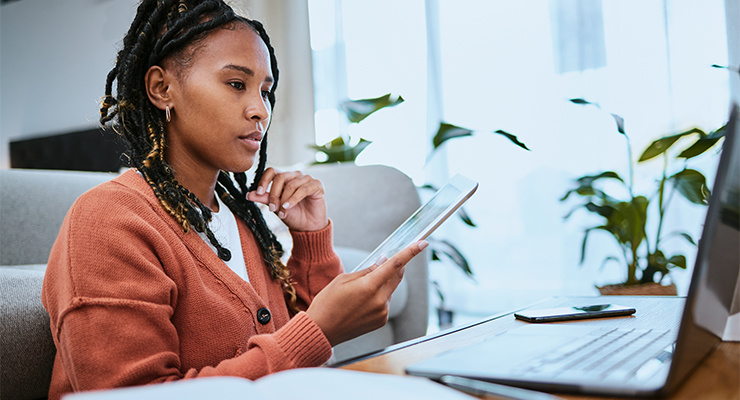 A woman holding a tablet while doing some research on a laptop.