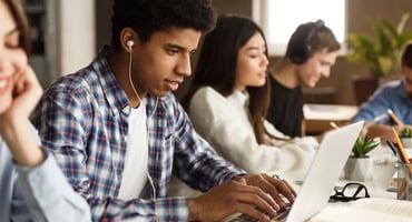 A male student sitting at a table with other students while checking his application status on a laptop