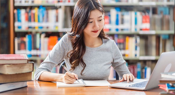 A female student sitting at a table in the library working on college decisions on her laptop