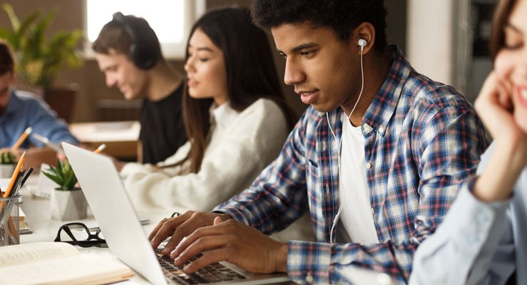 A male student wearing headphones reads a college decision email on his laptop at a table surrounded by other students