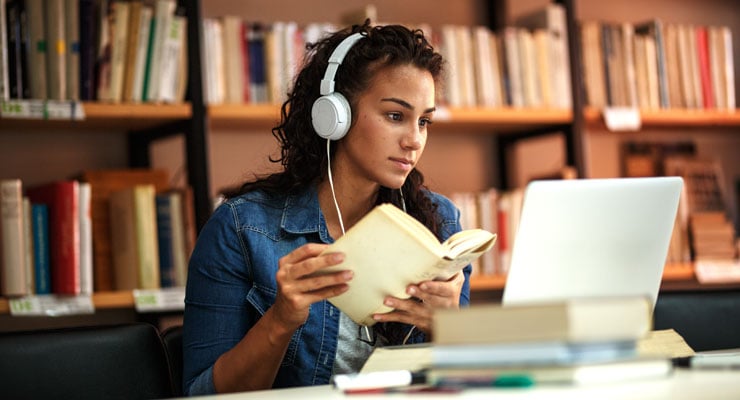 A college student studies in her school library while listening to music on her laptop with headphones