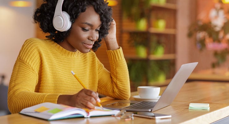 Female student wearing headphones and writing her notes down from her laptop learning online.