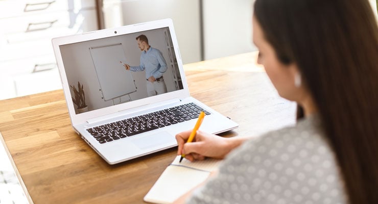 female student participating in online learning on her laptop at home taking notes from her online class.