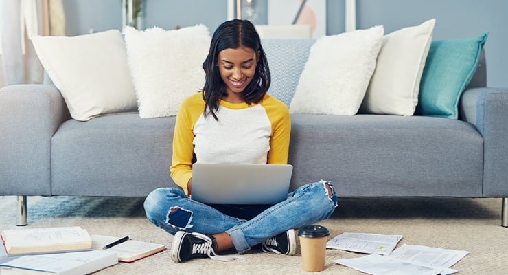 A student sitting by a couch and using her laptop.