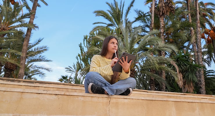 A student sitting cross-legged on campus with a tablet in her hand.