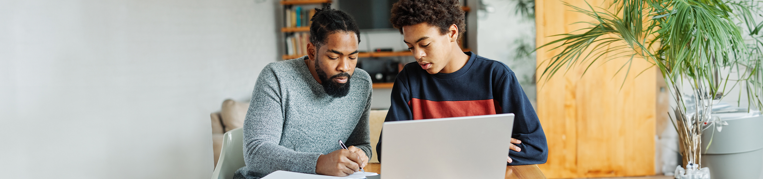 A father and his son taking notes on a piece of paper with a laptop open in front of them.