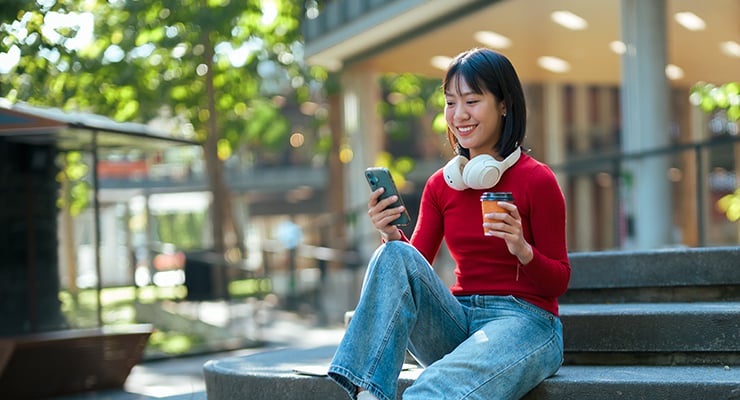 A student with headphones around her neck, drinking a cup of coffee, and scrolling on her phone.