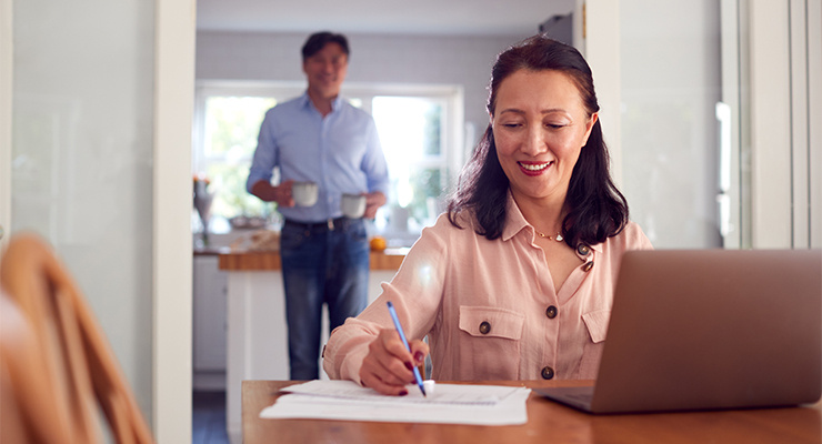 A mother signing documents with the father in the background with two cups of coffee.