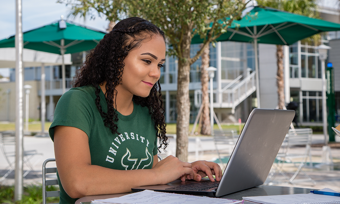 A female student using her laptop outside and taking advantage of the test fee waivers.