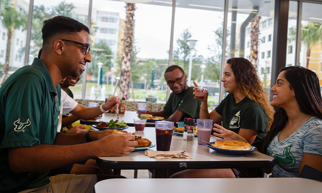 A group of students sitting and eating together in a dining hall, adjusting to the college life.